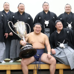 Sumo wrestler Hoshoryu sits with the Emperor's Cup after winning the 2025 hatsu basho (new year tournament). He's flanked by fellow sumo wrestlers Aku, Sadanoumi, Churanoumi, Meisei, Gonoyama, Hiradoumi and Ura of his ichimon (stable group).