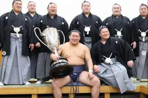 Sumo wrestler Hoshoryu sits with the Emperor's Cup after winning the 2025 hatsu basho (new year tournament). He's flanked by fellow sumo wrestlers Aku, Sadanoumi, Churanoumi, Meisei, Gonoyama, Hiradoumi and Ura of his ichimon (stable group).