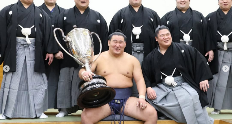 Sumo wrestler Hoshoryu sits with the Emperor's Cup after winning the 2025 hatsu basho (new year tournament). He's flanked by fellow sumo wrestlers Aku, Sadanoumi, Churanoumi, Meisei, Gonoyama, Hiradoumi and Ura of his ichimon (stable group).