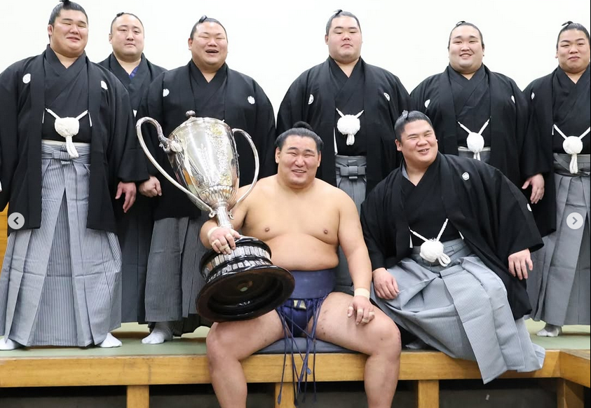 Sumo wrestler Hoshoryu sits with the Emperor's Cup after winning the 2025 hatsu basho (new year tournament). He's flanked by fellow sumo wrestlers Aku, Sadanoumi, Churanoumi, Meisei, Gonoyama, Hiradoumi and Ura of his ichimon (stable group).